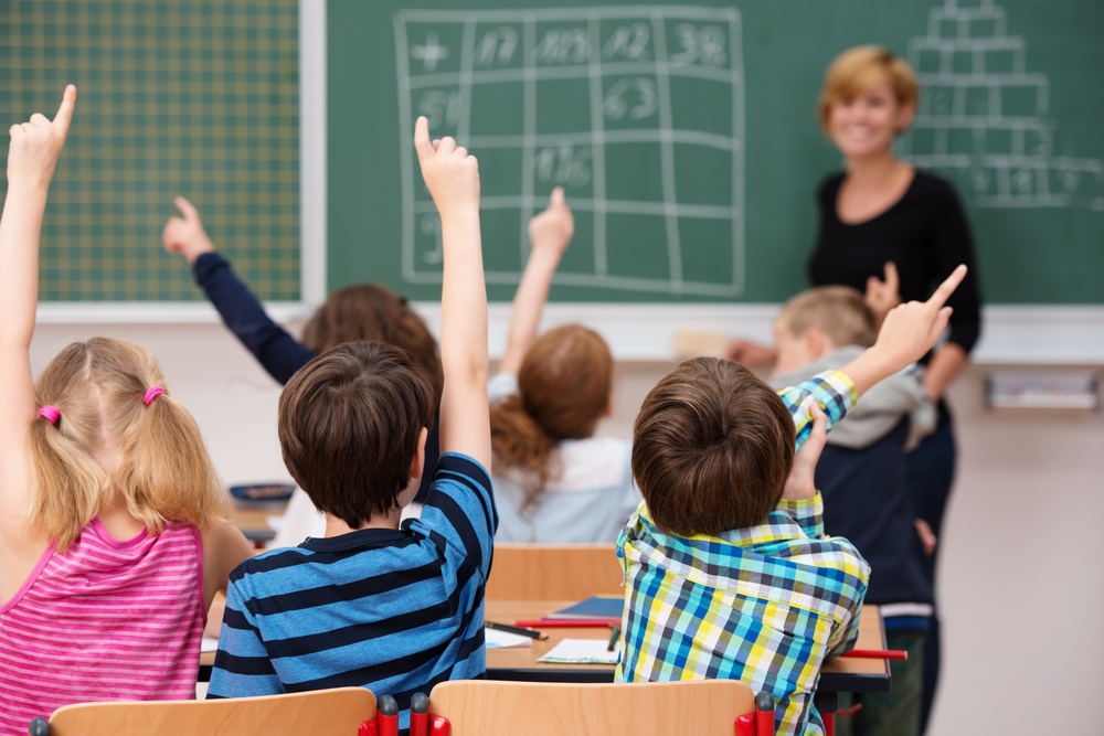 Intelligent group of young school children all raising their hands in the air to answer a question posed by the female teacher, view from behind.jpeg