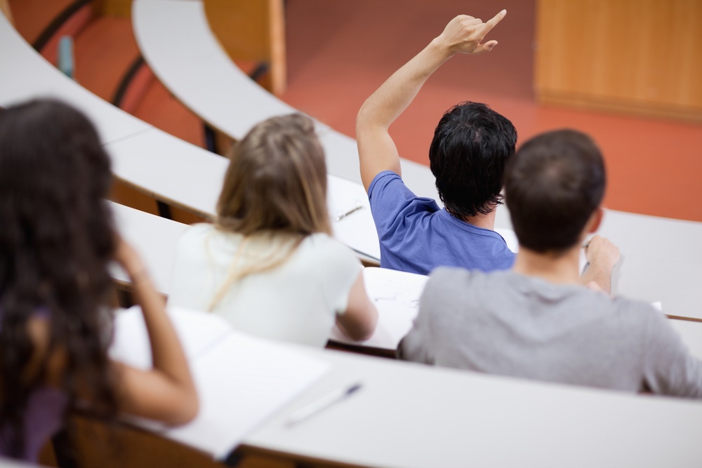 Jovem estudante levantando a mão para perguntar enquanto seus colegas tomam nota sobre a aula
